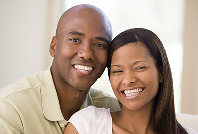 The image shows a man and woman posing closely together, smiling at the camera, with the man on the left wearing a light-colored shirt and the woman on the right in a dark top, both looking happy and content.